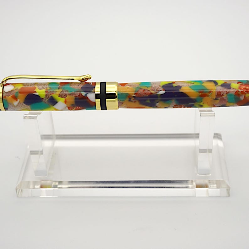 May include: A colorful, multi-colored, and patterned pen with gold accents and a black band. The pen is displayed on a clear acrylic stand.