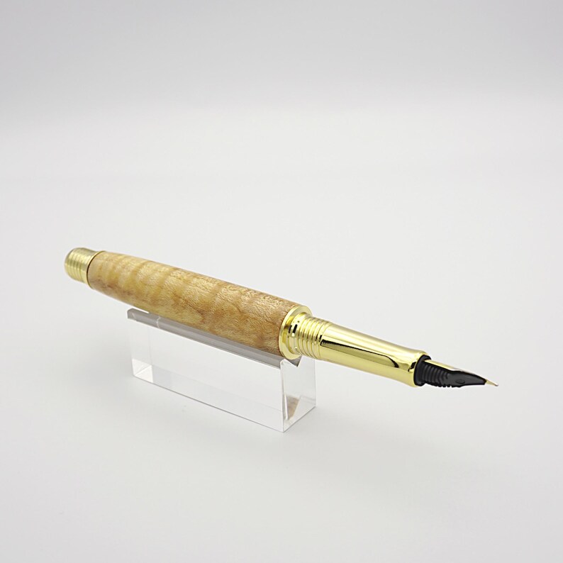 May include: A handcrafted fountain pen with a light brown, wood-grain barrel and gold-colored accents. The pen features a black nib and is displayed on a clear acrylic stand. A classic writing instrument.