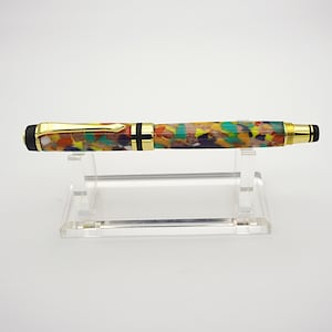 May include: A colorful, multi-colored, and patterned pen with gold accents and black end caps. The pen is displayed on a clear acrylic stand.