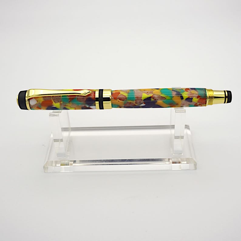 May include: A colorful, multi-colored, and patterned pen with gold accents and black end caps. The pen is displayed on a clear acrylic stand.