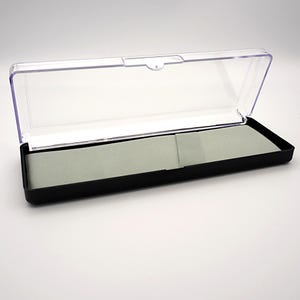 May include: A rectangular black pen case with a clear, hinged lid. The interior features a light gray felt lining. The case is open, revealing the inside. The case is designed to hold a pen or other small writing instrument.