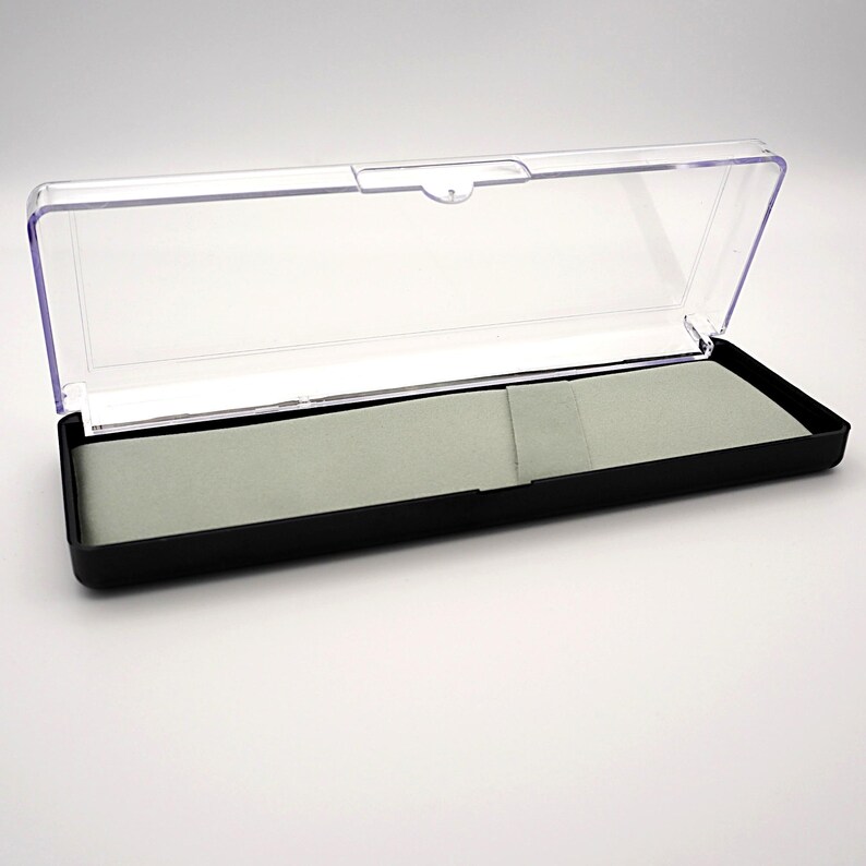 May include: A rectangular black pen case with a clear, hinged lid. The interior features a light gray felt lining. The case is open, revealing the inside. The case is designed to hold a pen or other small writing instrument.