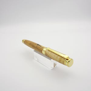 May include: A wooden pen with a light brown and tan finish, accented with gold-colored metal hardware. The pen is displayed on a clear acrylic stand, showcasing its design and craftsmanship. The pen's body has a natural wood grain pattern.