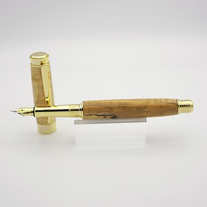 May include: A fountain pen with a light brown wooden barrel and gold-colored accents. The cap is off, revealing the nib. The pen rests on a clear acrylic stand, showcasing its design and craftsmanship.