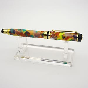 May include: A colorful, multi-colored, gold-trimmed pen with a black end cap, displayed on a clear acrylic stand.