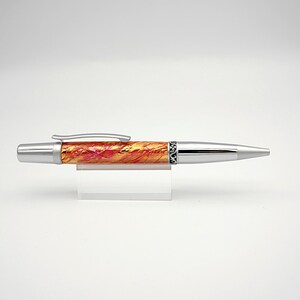 May include: A ballpoint pen featuring a barrel with a swirling orange, yellow, and pink marbled pattern. The pen has a polished silver-tone metal tip, a matching silver clip, and a decorative black band with intricate silver filigree detailing.