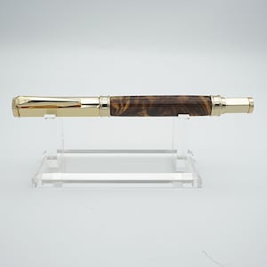 May include: A gold-plated fountain pen with a brown, wood-effect barrel. The pen is presented on a clear acrylic stand against a white backdrop. The pen features a cap and nib, and the barrel has a distinctive pattern.