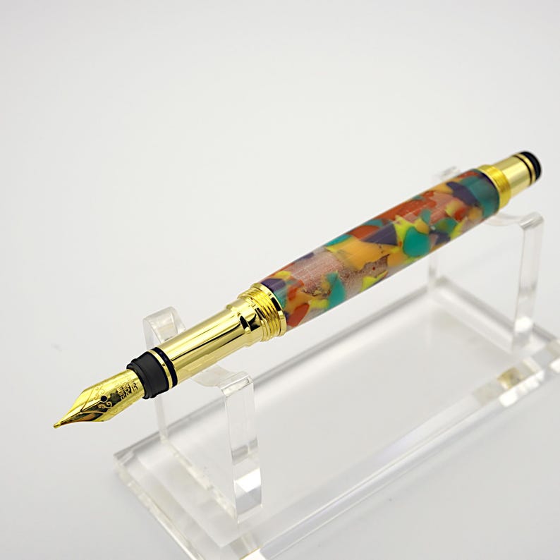 May include: A gold fountain pen with a colorful, marbled resin barrel. The pen is on a clear acrylic stand.
