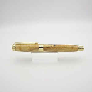 May include: A handcrafted wooden pen with gold-colored accents. The pen features a light brown, speckled barrel and a gold clip. The pen is displayed on a clear acrylic stand against a white background. A stylish writing instrument.