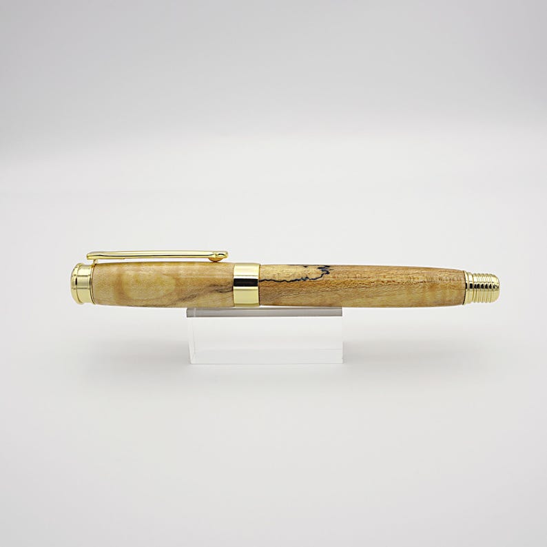 May include: A handcrafted wooden pen with gold-colored accents. The pen features a light brown, speckled barrel and a gold clip. The pen is displayed on a clear acrylic stand against a white background. A stylish writing instrument.