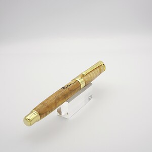 May include: A handcrafted wooden pen with a light brown and black marbled pattern. The pen features gold-colored accents on the clip, bands, and tip. The pen is displayed on a clear acrylic stand against a white background.