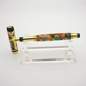 May include: A gold fountain pen with a colorful, multi-colored, abstract pattern on the barrel. The pen is on a clear acrylic stand.