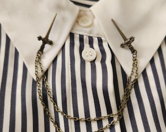 May include: A bronze-coloured collar pin set featuring two sword-shaped pins connected by a double chain. The pins are attached to a white collared shirt with navy blue stripes. The shirt has a button-down front.
