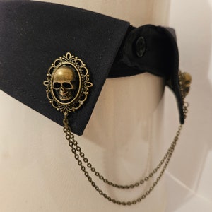 May include: A black fabric collar with a gold skull and chain detail. The skull is in an ornate frame and the chain is attached to the frame.