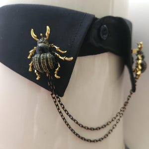 Könnte beinhalten: Ein schwarzer Hemdkragen mit einer dekorativen Käferbrosche. Die Brosche ist schwarz mit goldfarbenen Akzenten und kleinen, funkelnden Verzierungen. Eine zarte Kette hängt von der Brosche.