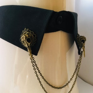 Puede incluir: Un cuello de tela negro con dos adornos de calavera y cadena de bronce. Las cadenas están conectadas y cuelgan del cuello.