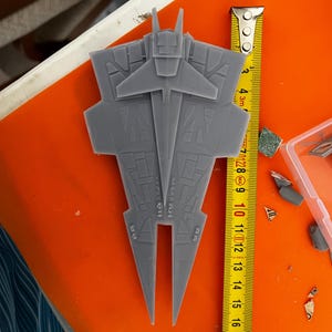 May include: A gray, detailed model of a spaceship, approximately 6 inches long, with wings and intricate designs. A yellow measuring tape is placed next to the model, showing the length. The model is on an orange surface.