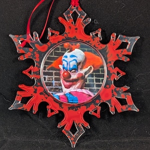 May include: A red snowflake ornament with a black and white image of a clown with a red nose and blue eyes. The ornament has a red and black splatter paint design.