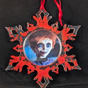 May include: A red and black snowflake-shaped Christmas ornament with a picture of a doll with red hair and blue eyes. The ornament has a red ribbon for hanging.