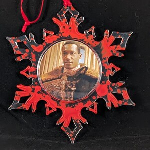 May include: A red and black snowflake ornament with a photo of a man in a fur coat inside. The snowflake is made of clear plastic and has a red ribbon attached.