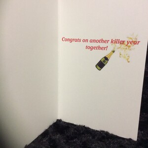 May include: A white card with the text "Congrats on another killer year together!" in red. A cartoon image of a champagne bottle popping with gold bubbles is to the right of the text.