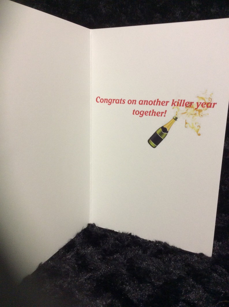 May include: A white card with the text "Congrats on another killer year together!" in red. A cartoon image of a champagne bottle popping with gold bubbles is to the right of the text.