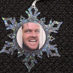 May include: A clear snowflake-shaped Christmas bauble with a glittery finish. The bauble features a round photo of a man with a surprised expression.