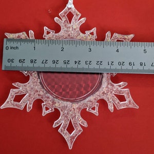 May include: A clear plastic snowflake ornament with a red glitter center. The ornament has a white ribbon for hanging and measures approximately 3 inches in diameter.