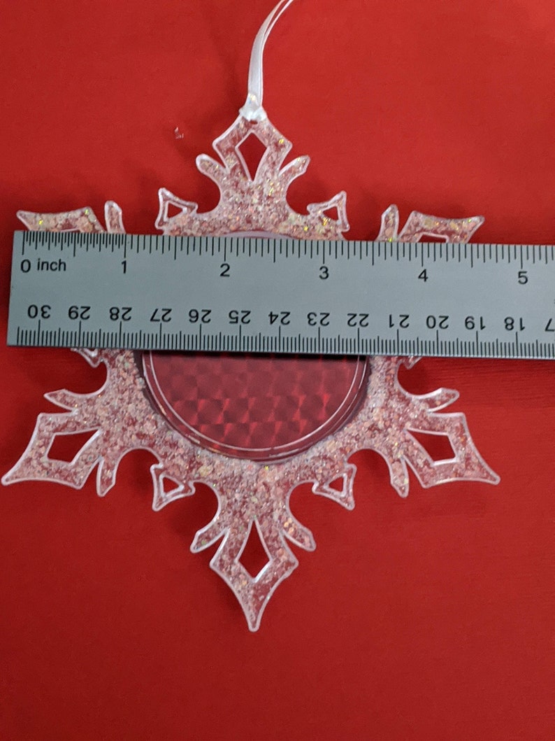 May include: A clear plastic snowflake ornament with a red glitter center. The ornament has a white ribbon for hanging and measures approximately 3 inches in diameter.