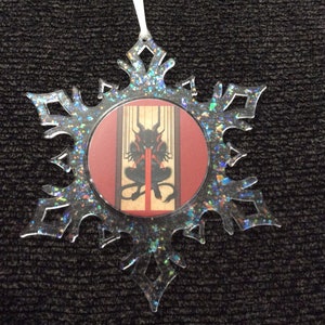 May include: A clear snowflake-shaped Christmas ornament with a red, white, and black image of a demon inside. The ornament is decorated with iridescent glitter.