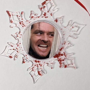 May include: A clear plastic snowflake ornament with red paint splatters and a round mirror in the centre. The mirror reflects a man's face with a sinister smile.