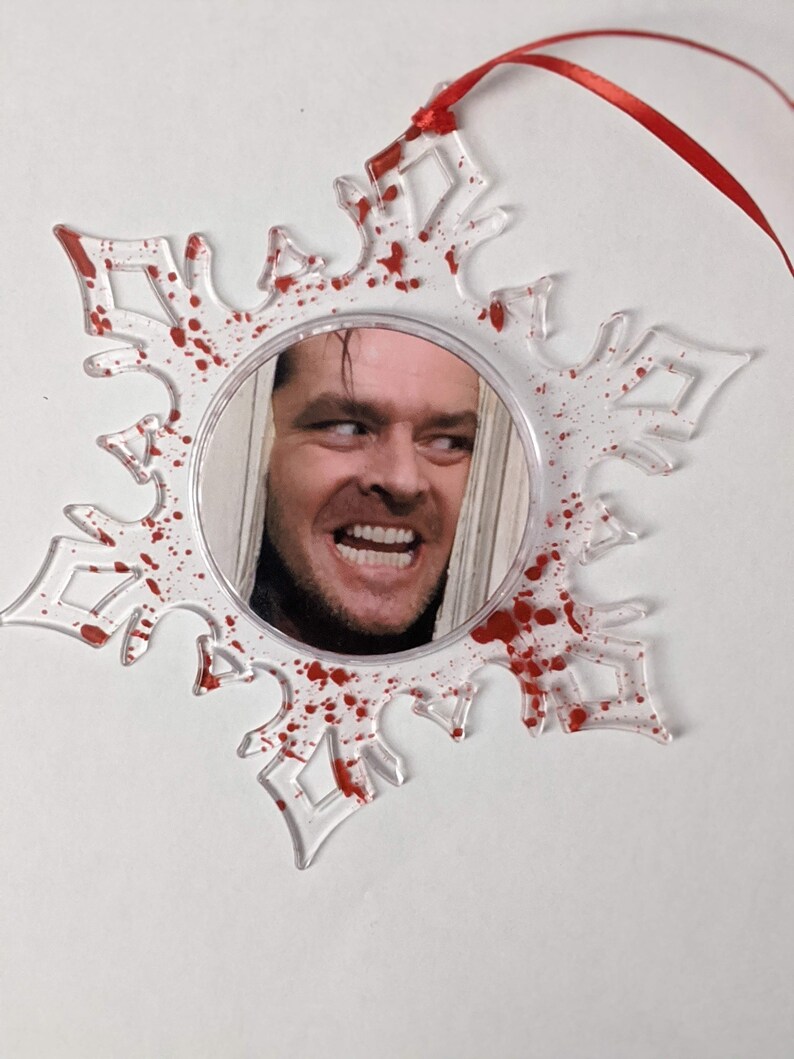 May include: A clear plastic snowflake ornament with red paint splatters and a round mirror in the centre. The mirror reflects a man's face with a sinister smile.