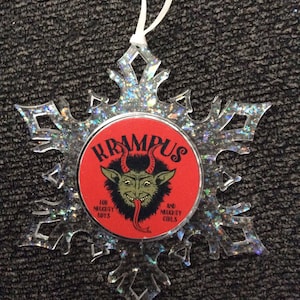 May include: A red and white snowflake ornament with a red circle in the center. The circle has a black and green illustration of a creature with horns and a long tongue. The text "Krampus" is written in black letters above the creature. The text "For Naughty Boys" is written in black letters on the left side of the creature and the text "And Naughty Girls" is written in black letters on the right side of the creature.