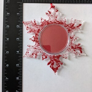 May include: A clear glass snowflake ornament with a red splatter paint design. The snowflake has a round opening in the center, which is filled with a red circle.