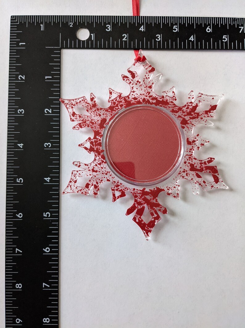 May include: A clear glass snowflake ornament with a red splatter paint design. The snowflake has a round opening in the center, which is filled with a red circle.