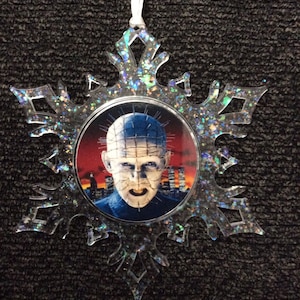 May include: A silver snowflake Christmas ornament with a round image of a character from the horror movie "Hellraiser" in the center. The character has a hooked face and is wearing a metal mask with spikes.