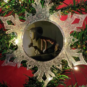 May include: A clear snowflake-shaped Christmas ornament with a glittery finish. The ornament features a circular image of a girl with a cat in a dark room.