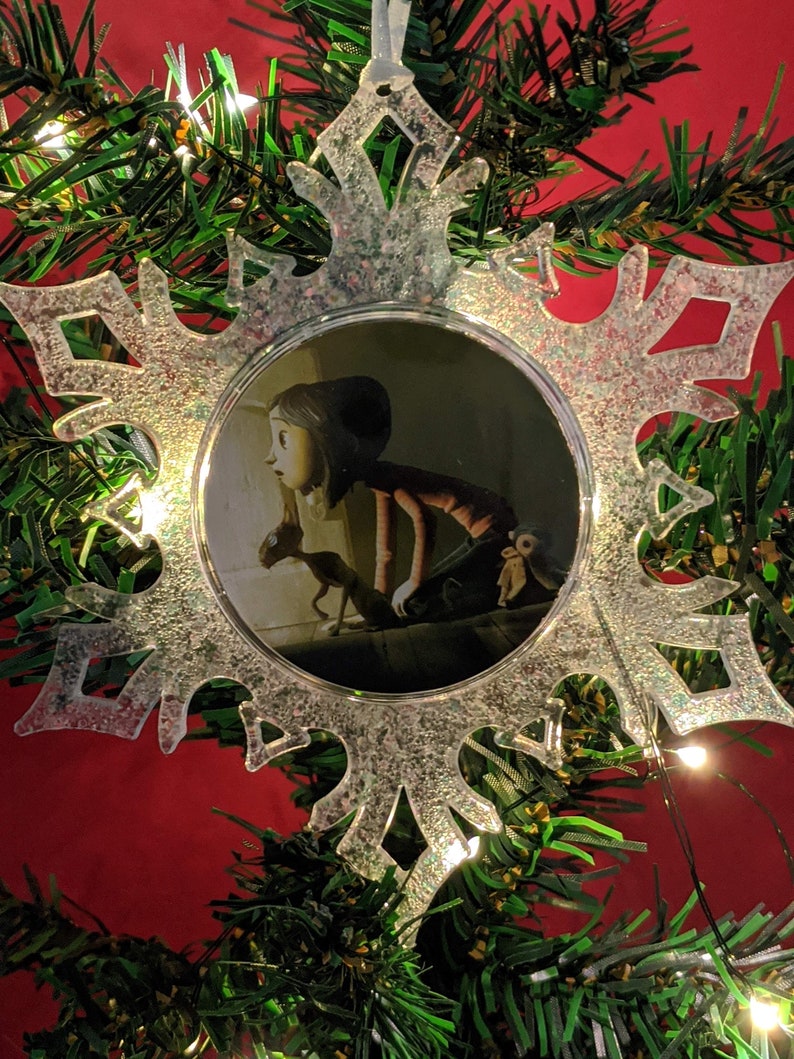 May include: A clear snowflake-shaped Christmas ornament with a glittery finish. The ornament features a circular image of a girl with a cat in a dark room.