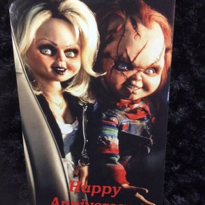 May include: A greeting card featuring a photo of the characters Chucky and Tiffany from the horror film Child's Play. The card has the text "Happy Anniversary" in red letters.