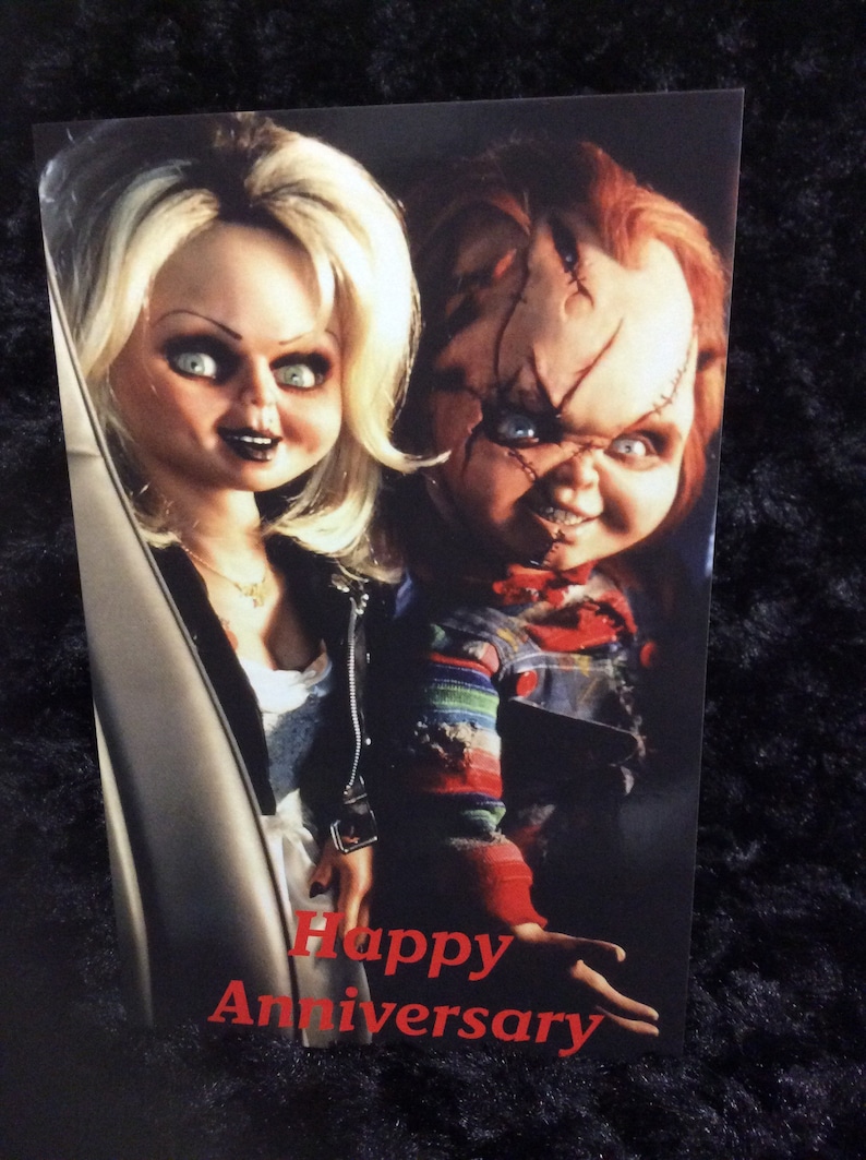 May include: A greeting card featuring a photo of the characters Chucky and Tiffany from the horror film Child's Play. The card has the text "Happy Anniversary" in red letters.