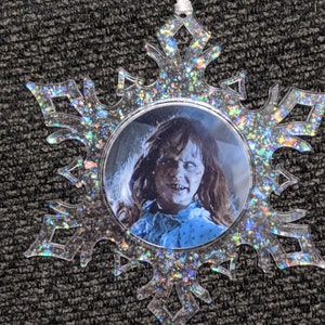 May include: A clear snowflake-shaped Christmas ornament with a glittery finish. The ornament features a round image of a girl with long dark hair, wearing a blue dress.