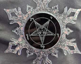 May include: A clear snowflake-shaped Christmas ornament with a black circle in the center. The circle has a pentagram with a goat's head inside and Hebrew lettering around the edge.