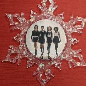 May include: A clear snowflake-shaped Christmas ornament with glitter embedded in the plastic. The ornament features a round image of four women in black clothing walking in a snowy setting.