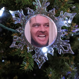 May include: A clear snowflake-shaped Christmas bauble with a glittery finish. The bauble features a photo of a man with a sinister smile.