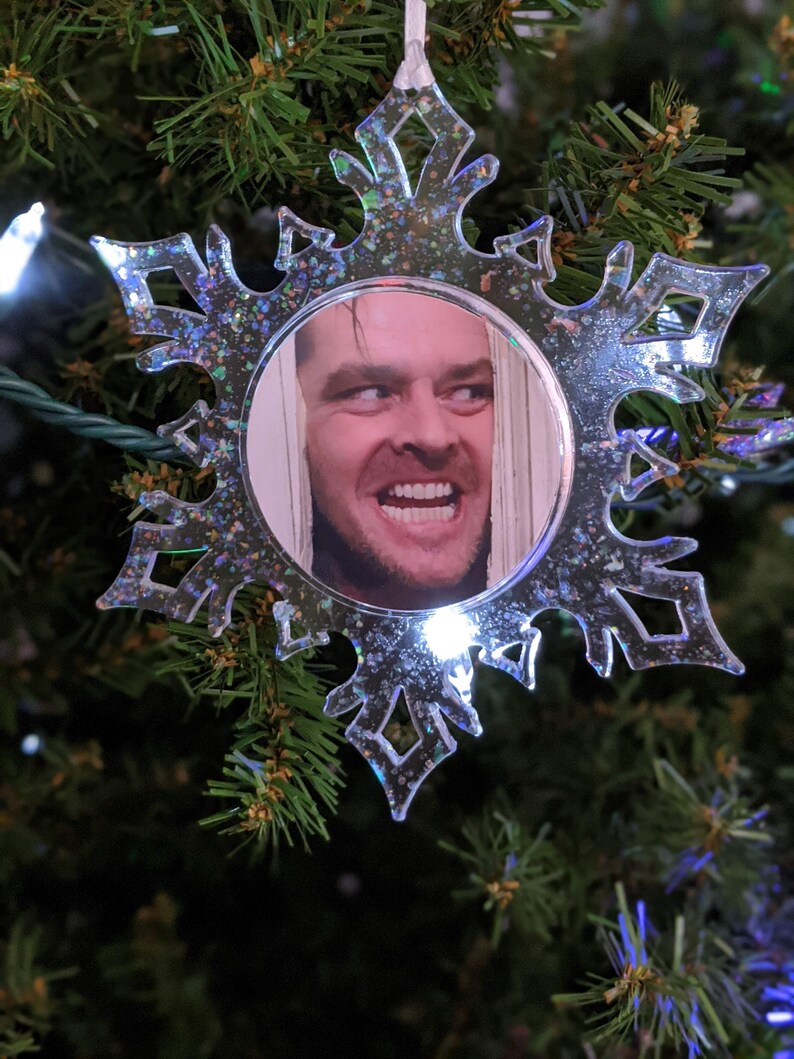 May include: A clear snowflake-shaped Christmas bauble with a glittery finish. The bauble features a photo of a man with a sinister smile.