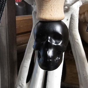 May include: A black skull-shaped car air freshener with a light brown wooden top, suspended by a black cord. The skull has a glossy finish. The background includes a white bone structure.