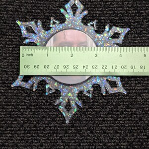 May include: A clear plastic snowflake ornament with a silver glitter finish. The ornament has a round mirror in the centre. The ornament is 7.6 cm in diameter.