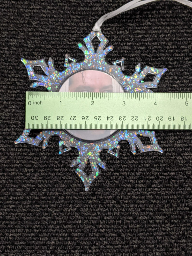 May include: A clear plastic snowflake ornament with a silver glitter finish. The ornament has a round mirror in the centre. The ornament is 7.6 cm in diameter.