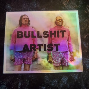 May include: A rectangular sticker featuring two people wearing pink turtlenecks and shorts. The text "BULLSHIT ARTIST" is printed in black across the image. The background has a rainbow-like effect.
