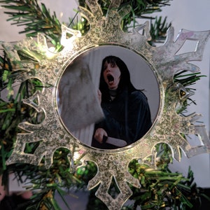 May include: A clear plastic snowflake ornament with a picture of a woman with dark hair and a white shirt, looking surprised and holding a knife. The ornament is decorated with glitter.